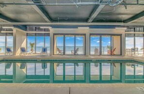 An indoor swimming pool with clear views of the ocean, lounge chairs, and a glass-enclosed area with a view.