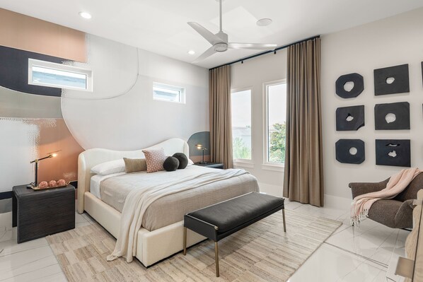 Un dormitorio moderno con una cama grande, un ventilador de techo, una mesita de noche con lámpara y vistas al exterior a través de una ventana.