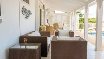 Uma área de estar coberta ao ar livre com móveis de vime e uma mesa de vidro, com vista para uma piscina.
