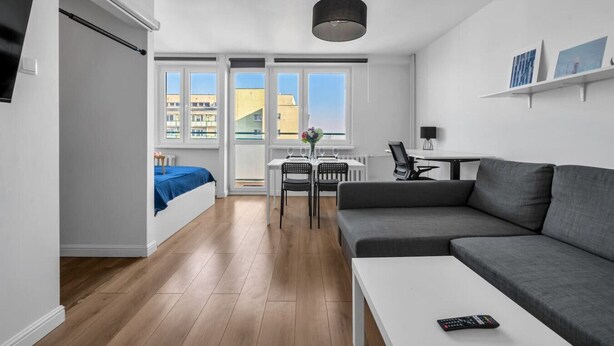 A modern, compact living space with a bed, a grey sofa, a dining table with chairs, and a small kitchen area. - Mokotów Studio- 14th Floor, Balcony - by Rentujemy (Warszawa)