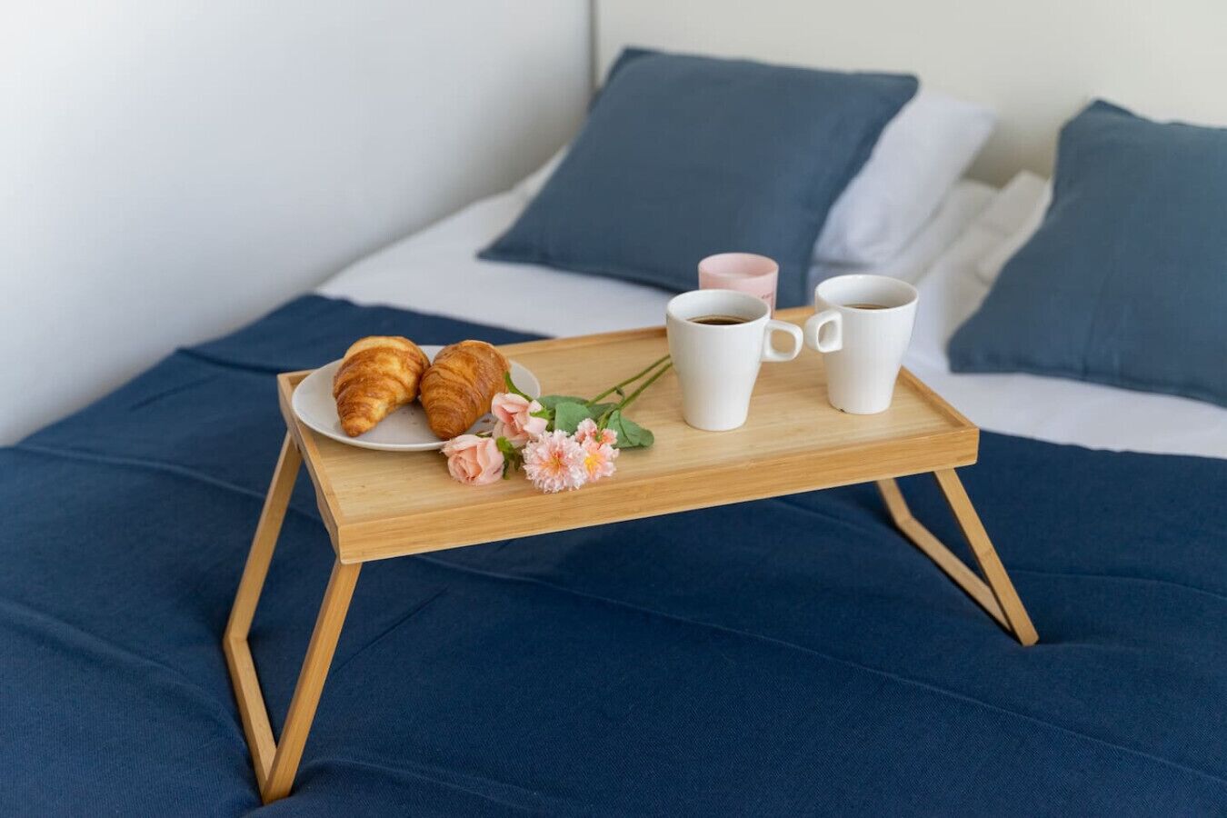 Een houten dienblad met croissants, koffiebekers en bloemen op een bed met blauw beddengoed en kussens.