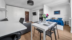 A modern hotel room with a dining area, a sofa, a bed with a blue cover, a TV mounted on the wall, and a flower vase on the dining table. - Mokotów Studio- 14th Floor, Balcony - by Rentujemy (Warszawa)