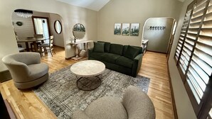 A spacious living room with a green sofa, a round coffee table, and a grey armchair. There is a dining area with a table and chairs in the background.