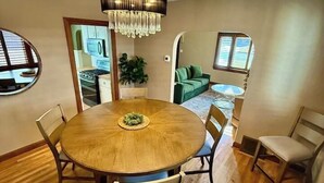 A dining area with a round wooden table, six chairs, a chandelier, and a view of a kitchen and living area through an archway.