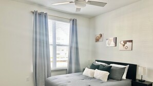Une chambre à coucher avec un lit, une table de chevet, un ventilateur de plafond et une fenêtre avec des rideaux.