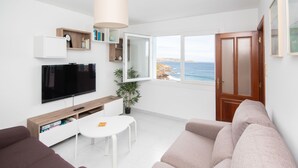 Un moderno salón con una televisión de pantalla plana, una mesita, un sofá y vistas al mar a través de un ventanal abierto.