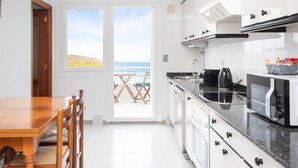 Una cocina con armarios blancos, encimera negra y vista a la playa a través de un ventanal corredero.