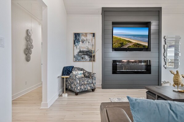 A modern living room with a flat-screen TV mounted on a grey wall, a patterned armchair, and a small table with a decorative turtle figurine. - Luxury Beachfront Home w/ Hot Tub & Games (Crystal City)