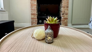 A living room with a brick fireplace, a round wooden table with a white decorative item, a red potted succulent, and a silver candle holder. - 4-bed 3 Bath House in Leicester City (Leicester)