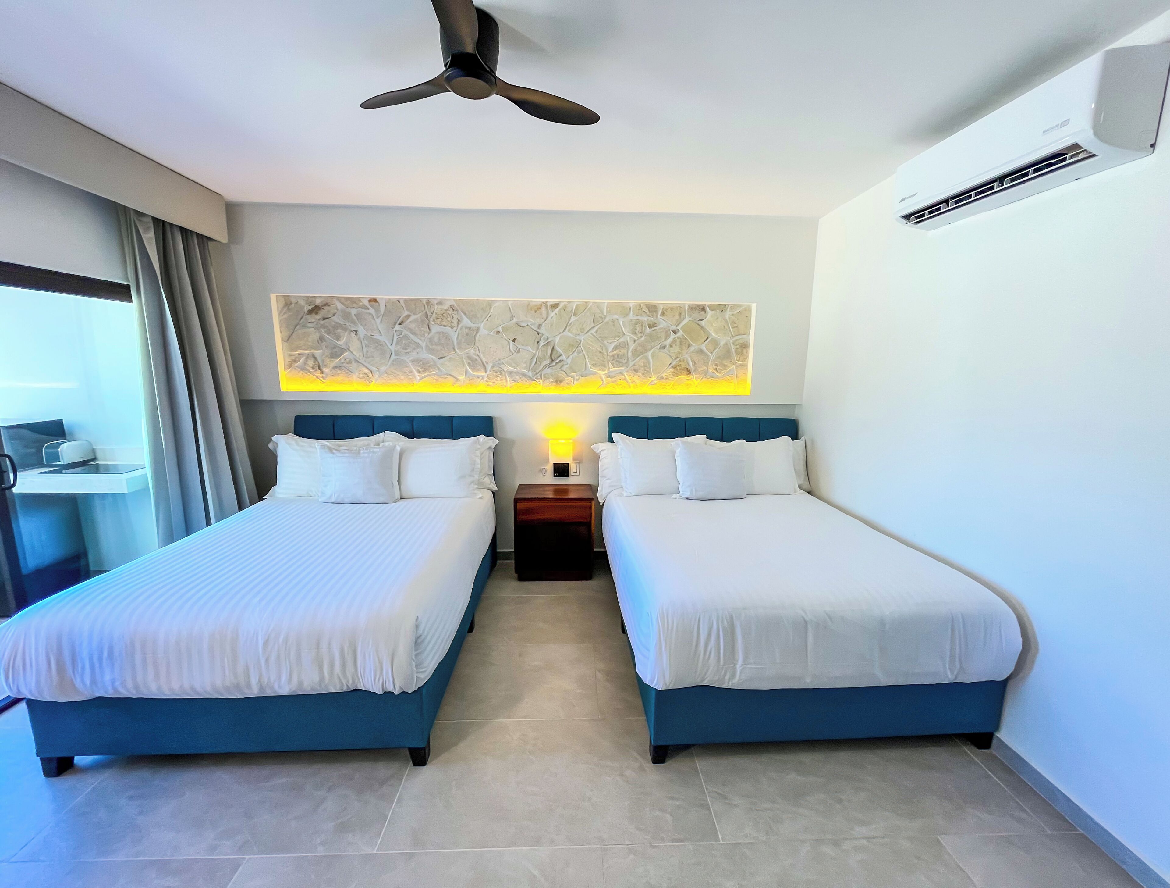 A hotel room with two beds, a ceiling fan, an air conditioning unit, a small table with a lamp, and a stone-patterned wall decoration.