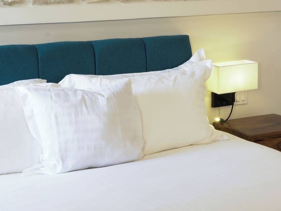 A neatly made bed with white linens and a teal headboard. A bedside lamp is on to the right, and a wooden nightstand is visible.