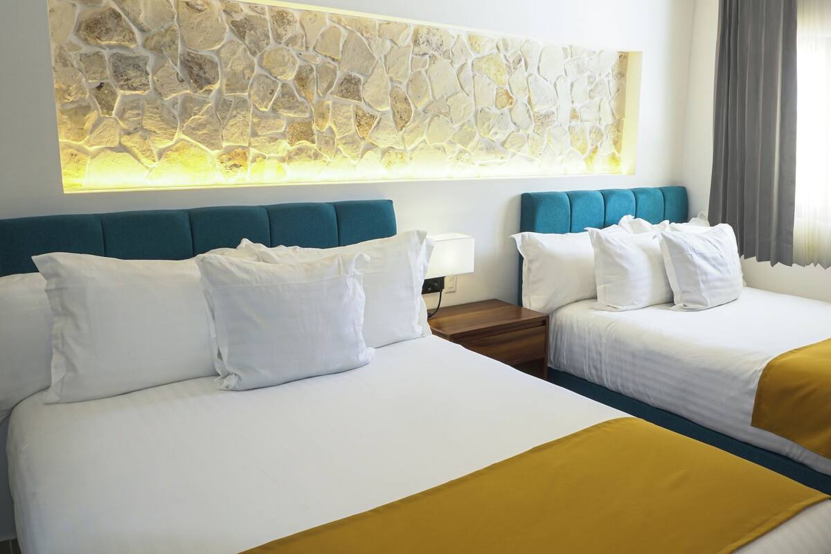 Ein Hotelzimmer mit zwei Betten, einem Kunstwerk aus Stein darüber, einem Nachttisch mit Lampe und einem Fenster mit Vorhängen.