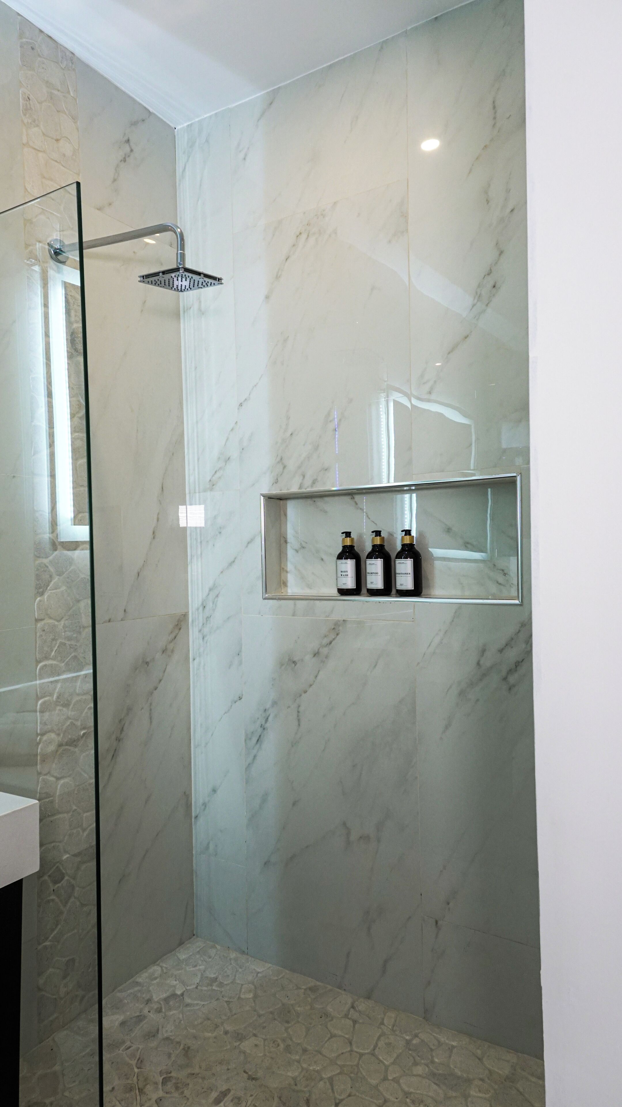 A modern bathroom with a glass shower enclosure, marble walls, and a shelf with three bottles.