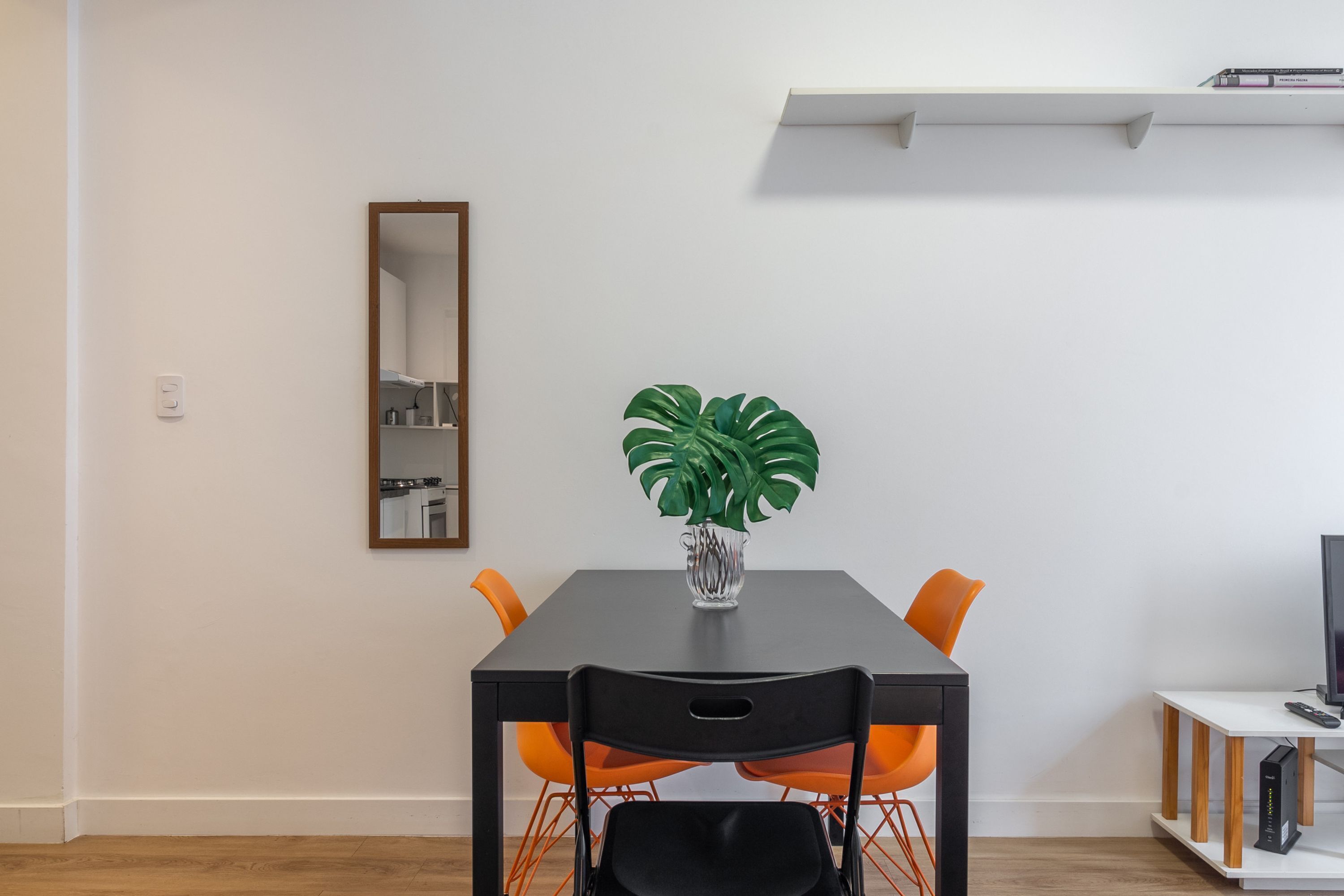 A dining area with a black table, orange chairs, a potted plant, a mirror, and a white shelf above the table.