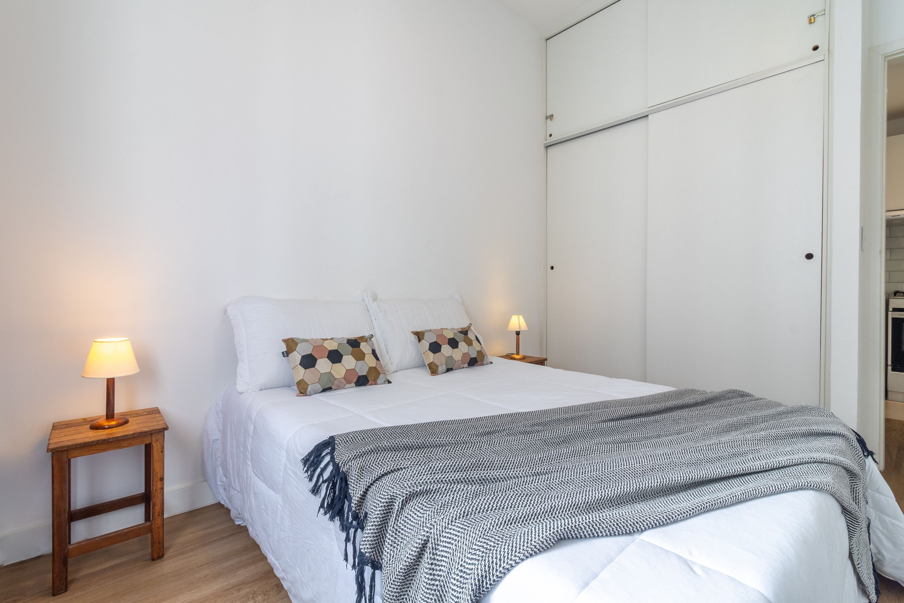 A neatly made bed with a patterned throw, two bedside lamps, and a white wardrobe in the background.
