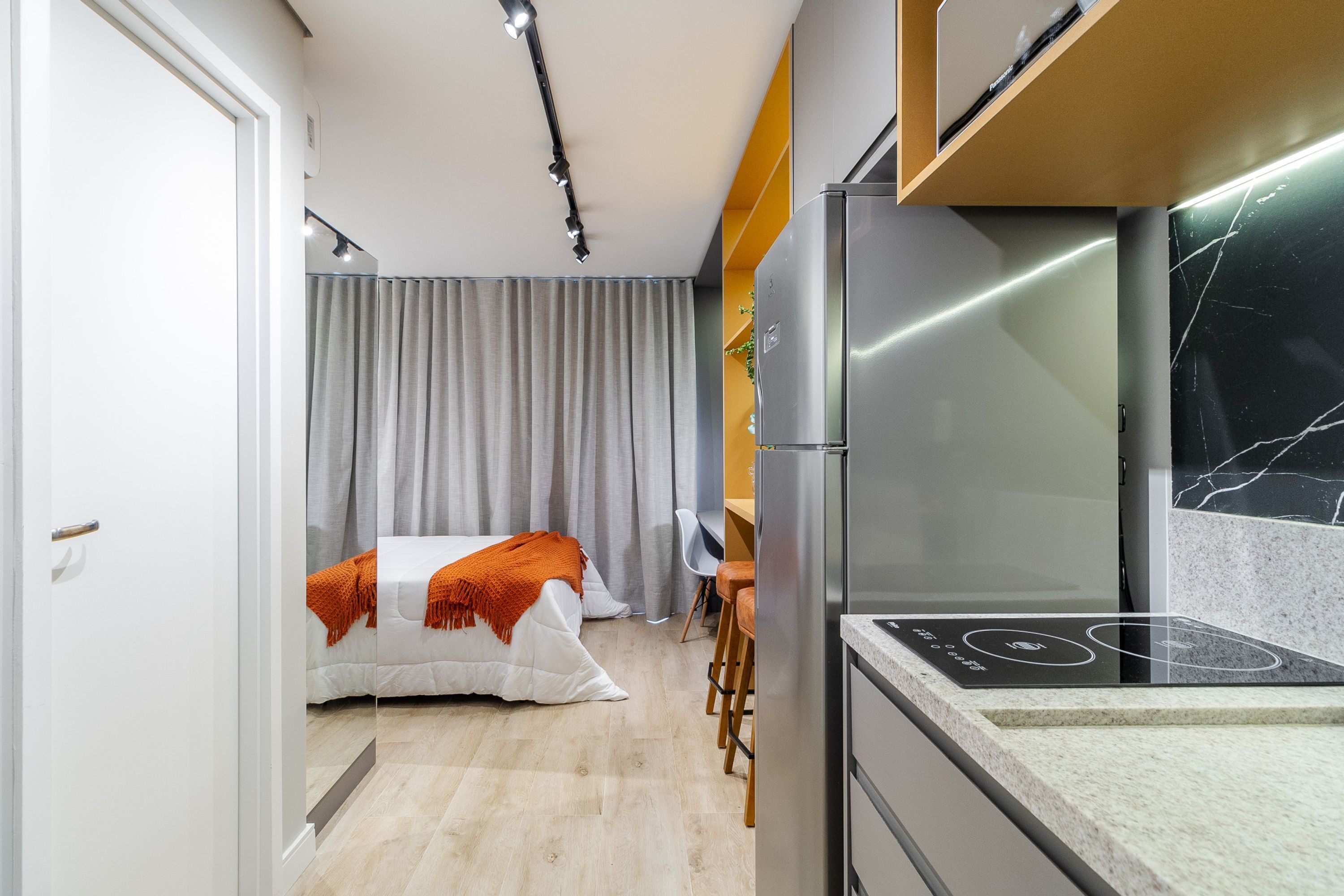 A compact hotel room with a bed, a desk, a chair, a refrigerator, and a kitchen area with a stove.