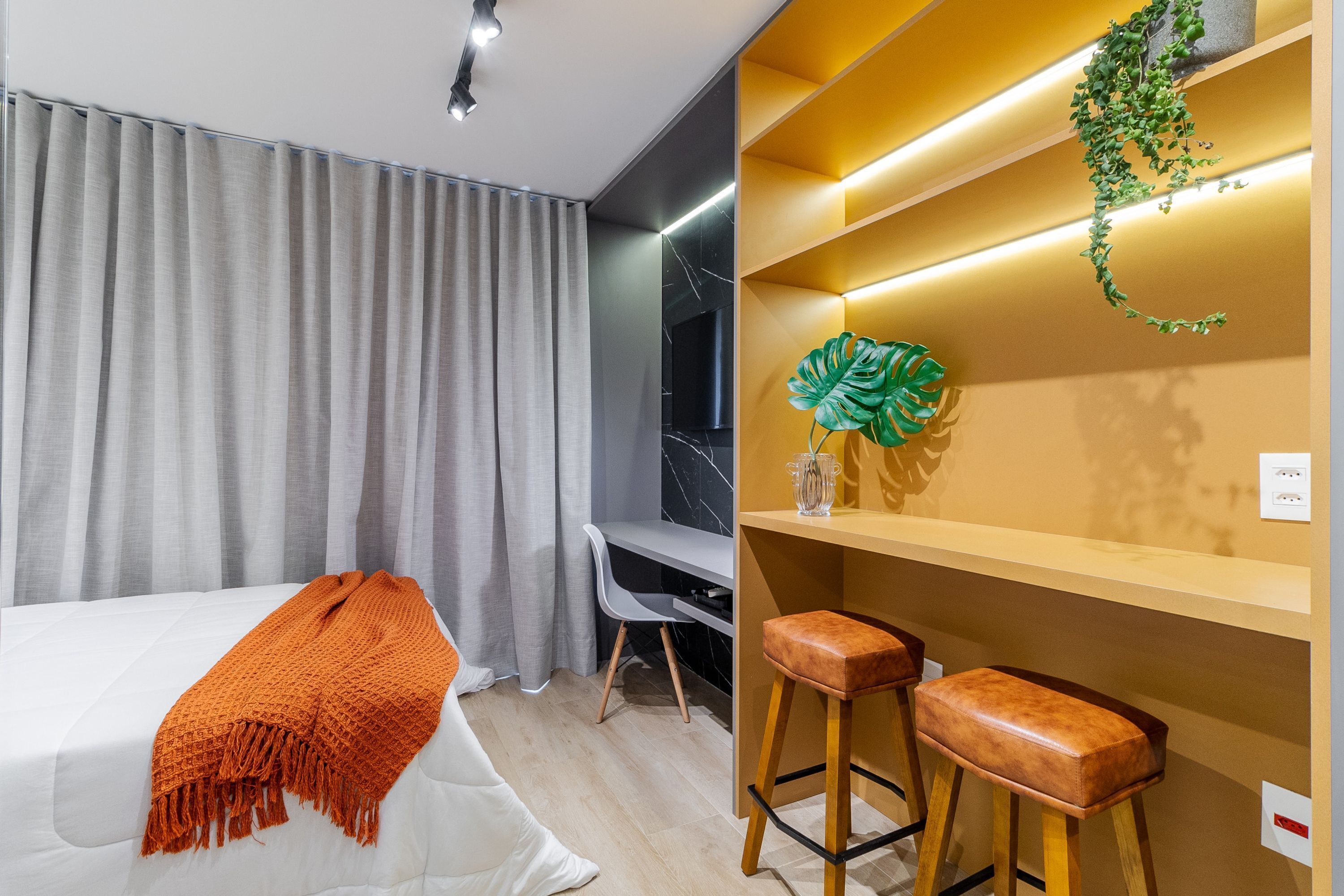 A modern hotel room with a bed, a desk, two bar stools, and a shelf with decorative items.