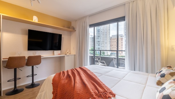 A hotel room with a bed, a TV, two bar stools, a balcony with a view of buildings, and a window with curtains. - Mingodoy in S o Paulo (São Paulo)
