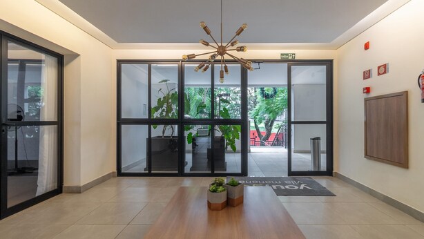 A modern lobby with a wooden reception desk, a unique chandelier, large glass doors leading to an outdoor area with plants, and a "Welcome" mat. - Machado 238 Ap 137 in S o Paulo (São Paulo)