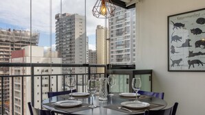 A dining area with a table set for four, surrounded by chairs, with a cityscape view through large windows. - Machado 238 Ap 137 in S o Paulo (São Paulo)