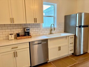 A kitchen with white cabinets, a stainless steel dishwasher, a refrigerator, a window, and a coffee maker on the counter.