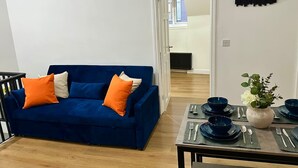 A living room with a blue sofa, orange and white cushions, a dining table set with blue plates and silverware, and a vase with flowers.