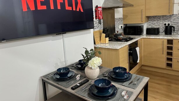 A modern kitchen with a flat-screen TV displaying "NETFLIX" in red letters, a dining table set with blue bowls and a vase of flowers, and wooden cabinets.