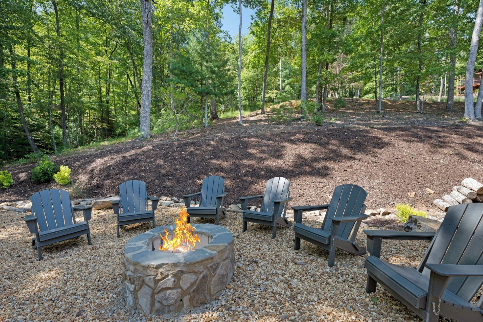 A fire pit surrounded by Adirondack chairs in a wooded outdoor area.