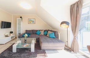 Ein modernes Wohnzimmer mit einem Ecksofa, einem Glastisch, einem Flachbildfernseher und einem großen Fenster mit Vorhängen.