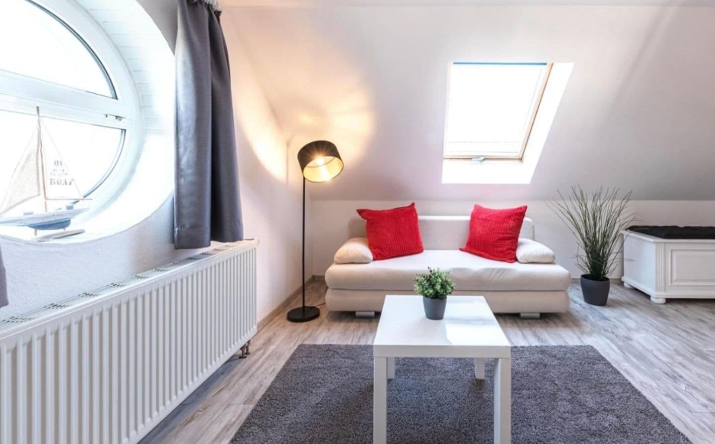 A modern, well-lit living room with a white sofa, red cushions, a small table with a plant, a floor lamp, and a window with a view of a sailboat.