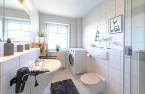 Un baño moderno con un lavamanos grande, inodoro y lavadora. Hay plantas en macetas y una ventana pequeña que proporciona luz natural.
