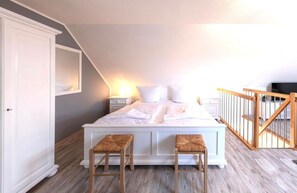 Un dormitorio con techo inclinado, una cama con ropa de cama blanca, dos taburetes de madera, un armario blanco y una barandilla de madera.