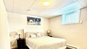 Une chambre à coucher comprenant un lit, deux tables de chevet, une commode, un tapis à motifs, un luminaire au plafond, une fenêtre et un tableau accroché au mur.