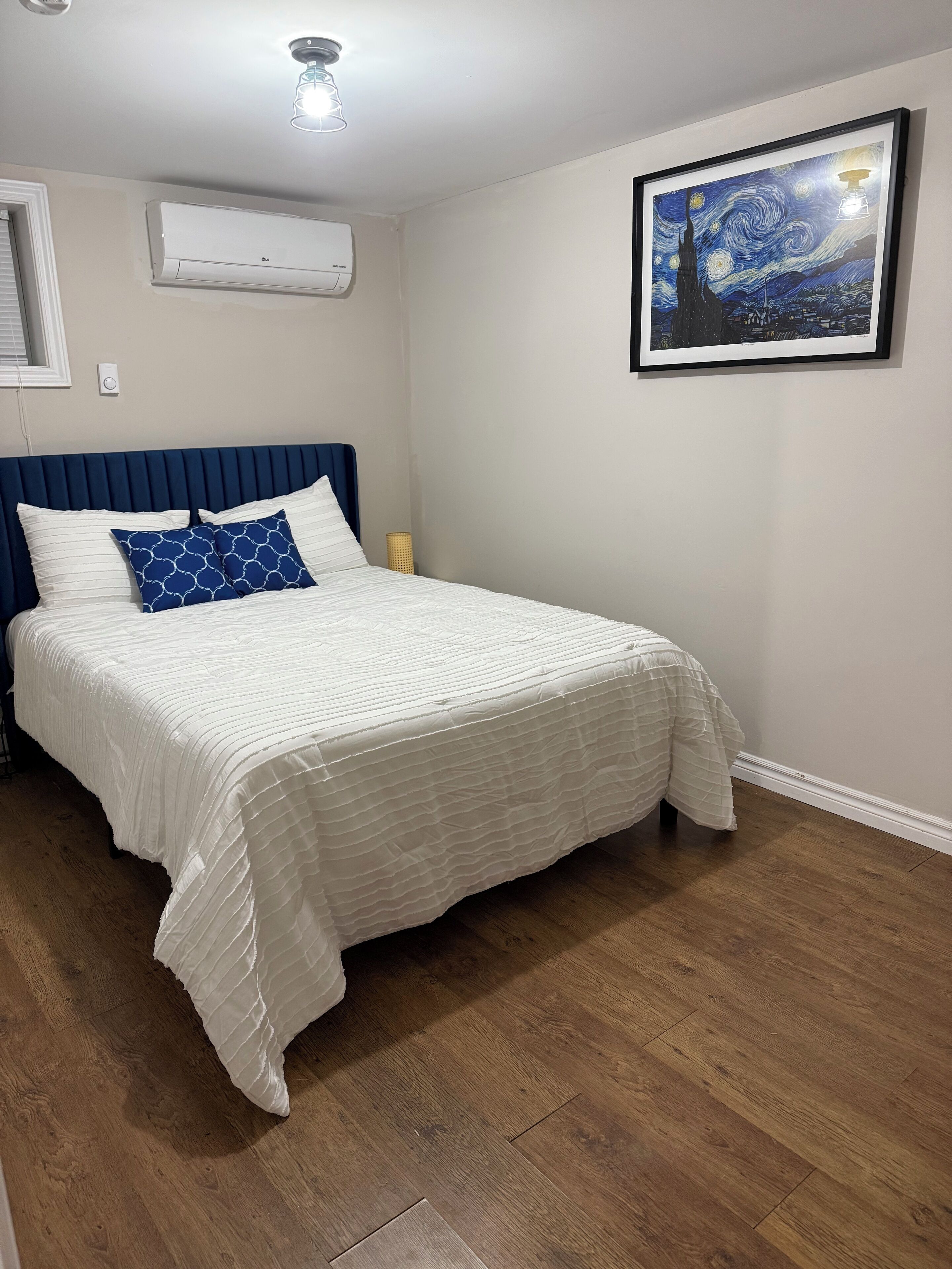 A bedroom with a bed, a ceiling light, an air conditioner, a framed artwork, and a wooden floor.