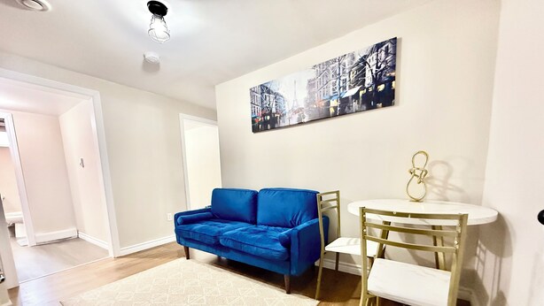 Un salon avec un canapé bleu, un tapis blanc, une table ronde avec une chaise, et un tableau représentant un paysage urbain accroché au mur.
