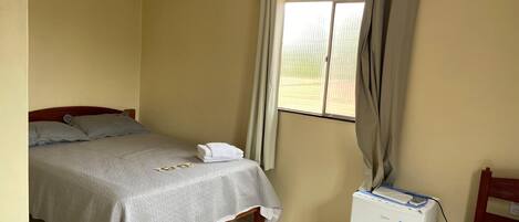 A small, clean room with a bed, a small table with a refrigerator, a window with curtains, and a tiled floor.
