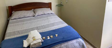 Una cama bien tendida con una manta azul y una toalla doblada, una mesita de noche con repisa de cristal, e interruptor de luz fijado en la pared.