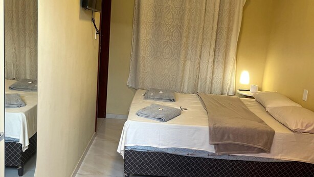 A bedroom with a bed, a bedside table, a mirror, a wall-mounted air conditioner, and a curtain. - Pousada BG Lençóis (Barreirinhas)