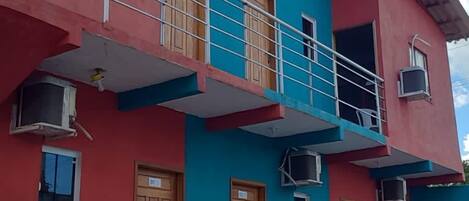 A two-story building with a red and blue facade, balconies with railings, and a small garden area with potted plants.