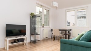 Een moderne woonkamer met een flatscreen televisie op een witte standaard, een glazen kast met decoratieve objecten, een eethoek met een tafel en stoelen, en een groene bank.