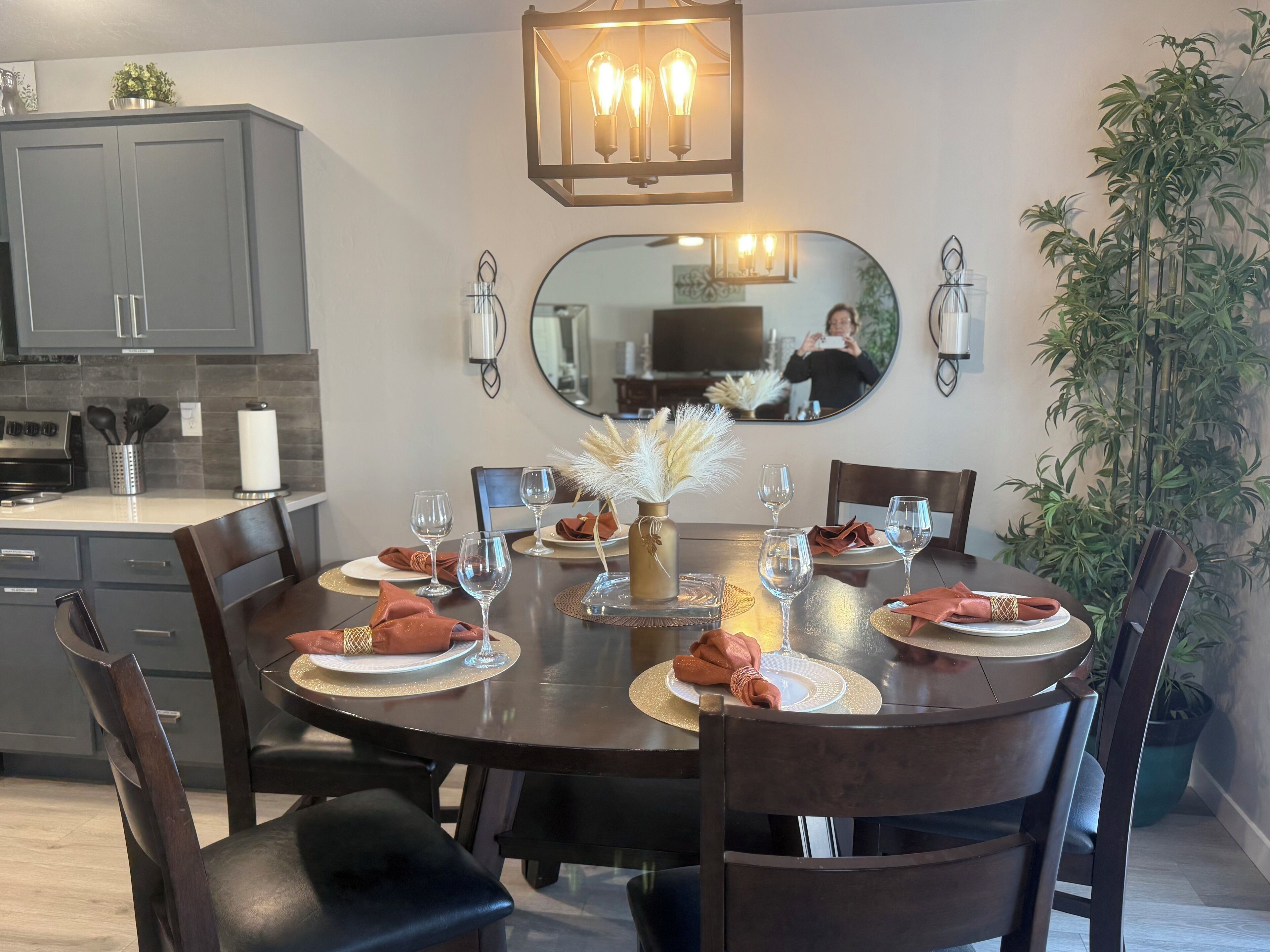Un coin repas est aménagé avec une table ronde pour six personnes, entourée de chaises en bois. À gauche, on trouve un espace cuisine avec des meubles gris et un four encastré. Un grand miroir est accroché au mur au-dessus de la table, et il reflète une partie de la pièce.