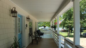 A covered porch with wooden flooring, white railings, and a blue door. There are several chairs and a potted plant on the porch.
