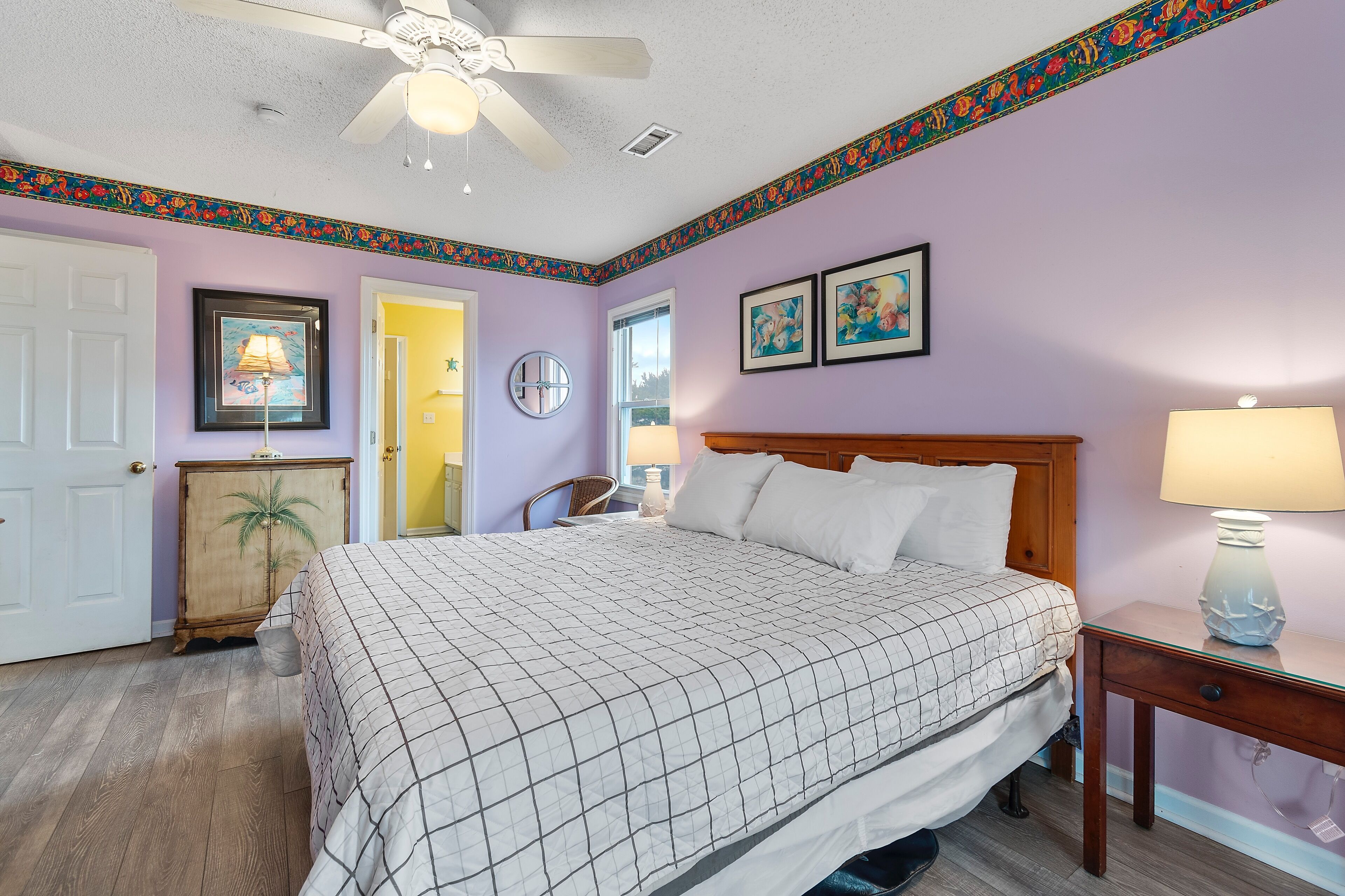 A bedroom with a bed, a nightstand, a lamp, a ceiling fan, a door, and a wall with a decorative border.
