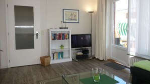 A living room with a bookshelf, a television, and a window with curtains. - Haus Teunis - Vacation in the best location, in the pedestrian zone in Wyk - Teunis (Wyk auf Föhr)