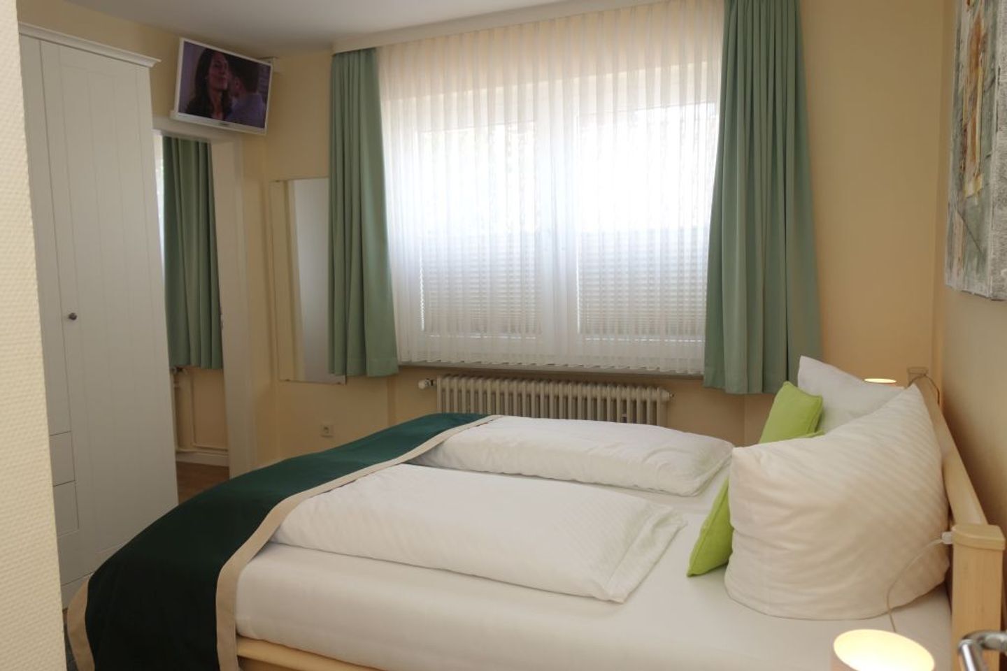 A hotel room with a bed, a radiator, a television on the wall, and a window with curtains.