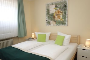 A double bed with white and green pillows, a bedside table with a lamp, and a wall-mounted abstract painting.