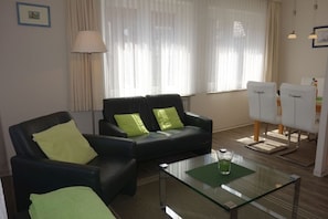 A living room with a glass coffee table, two black leather sofas with green cushions, a lamp, and a dining area with white chairs.