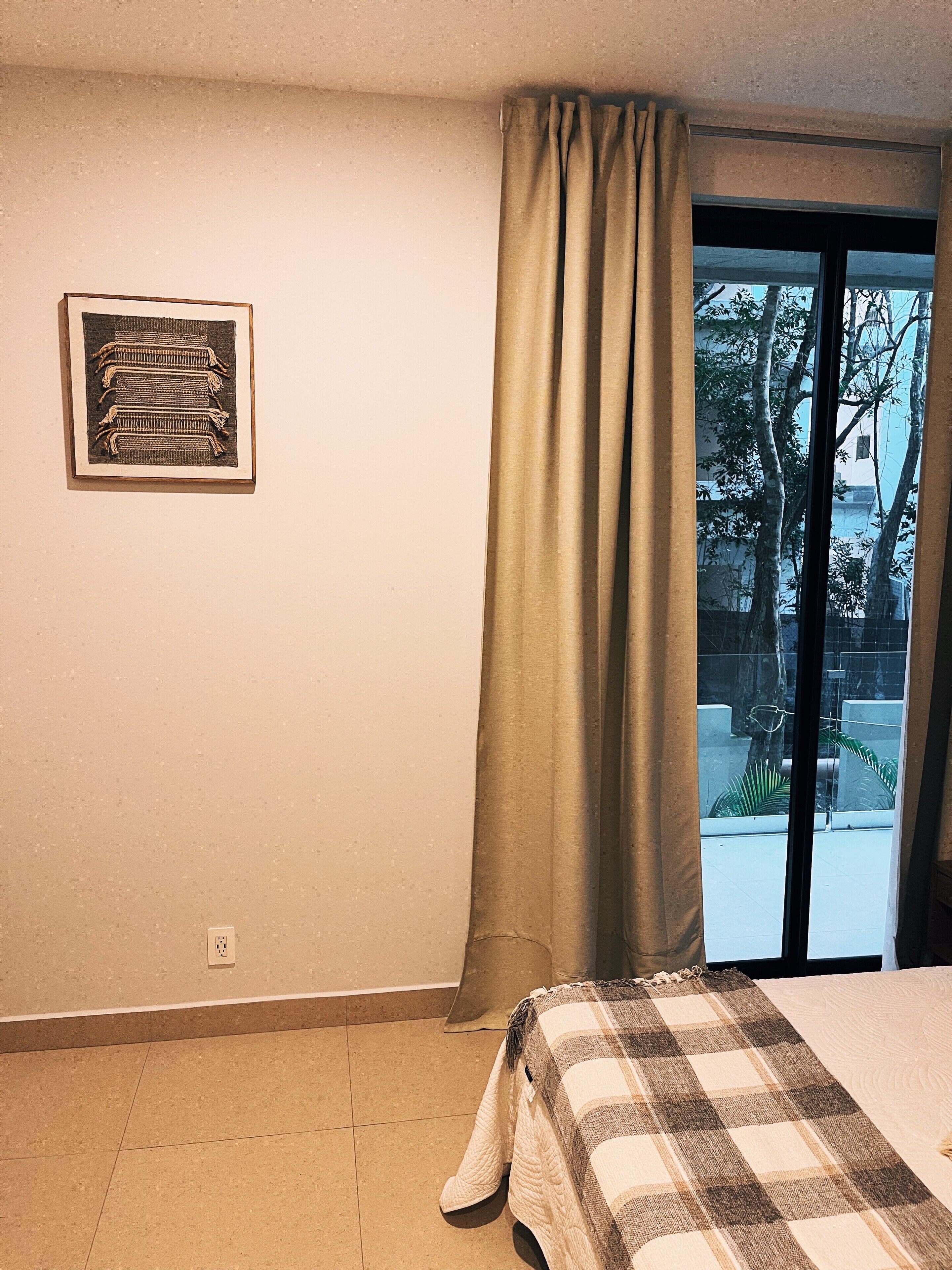 Een slaapkamer met een bed, een raam met gordijnen, een wandcontactdoos en een ingelijst schilderij.