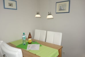 A dining area with a wooden table, two white chairs, a green tablecloth, two hanging pendant lights, and two framed pictures on the wall.