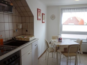 Una cocina con estufa, pileta y muebles de cocina, una mesa de comedor con sillas y una ventana que da a un edificio rojo.